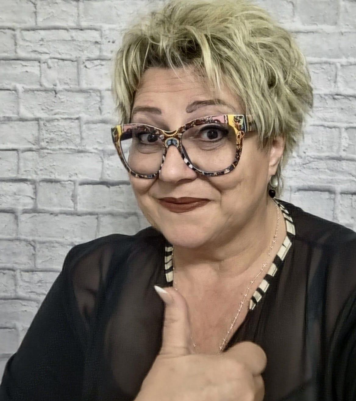 Eine Frau mit kurzen blonden Haaren, die eine Brille und ein schwarzes Hemd trägt, gibt einen Daumen hoch. Sie hat eine Halskette an und steht vor einer weißen Ziegelwand.