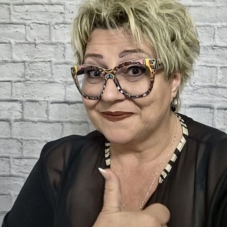 Eine Frau mit kurzen blonden Haaren, die eine Brille und ein schwarzes Hemd trägt, gibt einen Daumen hoch. Sie hat eine Halskette an und steht vor einer weißen Ziegelwand.