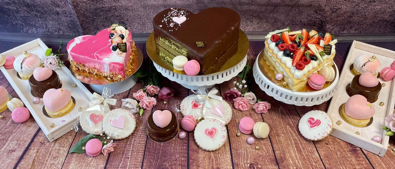 Eine Auswahl an herzförmigen Kuchen, Macarons und Keksen auf einem hölzernen Tisch, geschmückt mit rosa Blumen und Blütenblättern. Jeder Kuchen ist einzigartig verziert, einschließlich Schokoladen- und Erdbeerelemente.