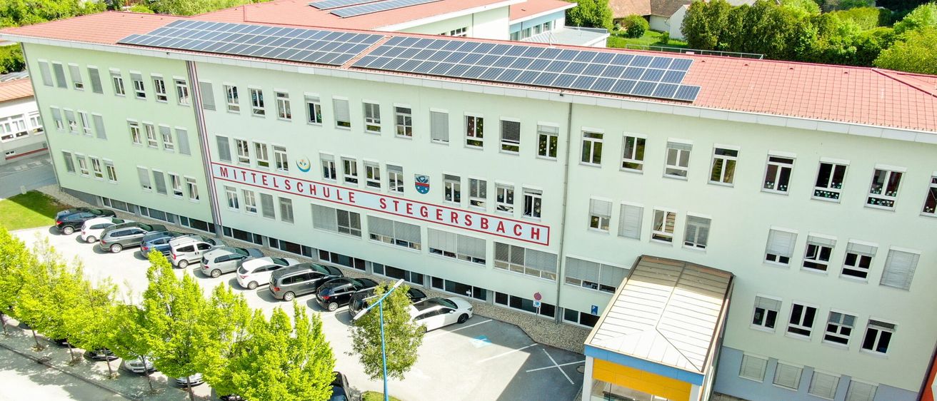 Luftaufnahme eines Schulgebäudes mit Solarpanelen auf dem Dach und einem Parkplatz mit mehreren Autos. Der Name der Schule ist Mittelschule Stegersbach.