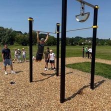 Ein Mann macht Klimmzüge an Outdoor-Trainingsstangen. Ein paar Leute beobachten. Bäume und eine Grasfläche sind im Hintergrund.