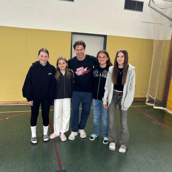 Fünf Personen, drei Mädchen und ein Mann, posieren für ein Foto in einer Turnhalle. Die Mädchen tragen Jacken und Sneakers. Der Mann trägt ein schwarzes T-Shirt mit einem Ninja Warrior-Logo.