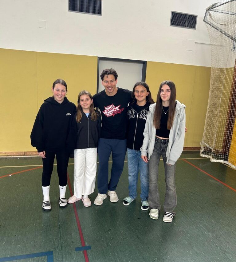 Fünf Personen, drei Mädchen und ein Mann, posieren für ein Foto in einer Turnhalle. Die Mädchen tragen Jacken und Sneakers. Der Mann trägt ein schwarzes T-Shirt mit einem Ninja Warrior-Logo.