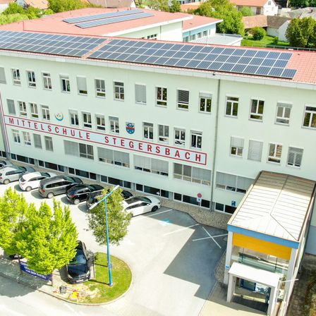 Eine Luftaufnahme der Mittelschule Stegersbach mit Solarpanelen, geparkten Autos und Bäumen. Das Gebäude hat viele Fenster.