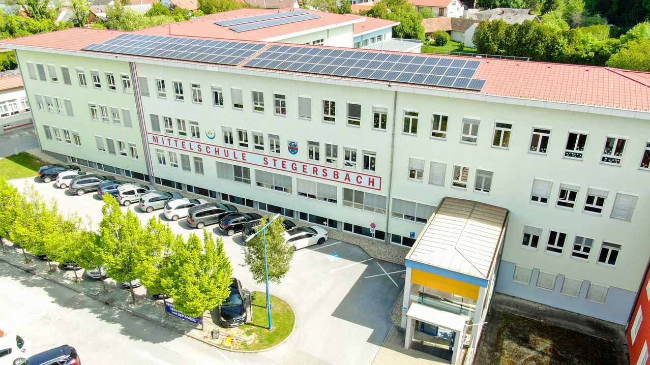 Eine Luftaufnahme der Mittelschule Stegersbach mit Solarpanelen, geparkten Autos und Bäumen. Das Gebäude hat viele Fenster.