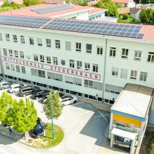 Eine Luftaufnahme der Mittelschule Stegersbach mit Solarpanelen, geparkten Autos und Bäumen. Das Gebäude hat viele Fenster.