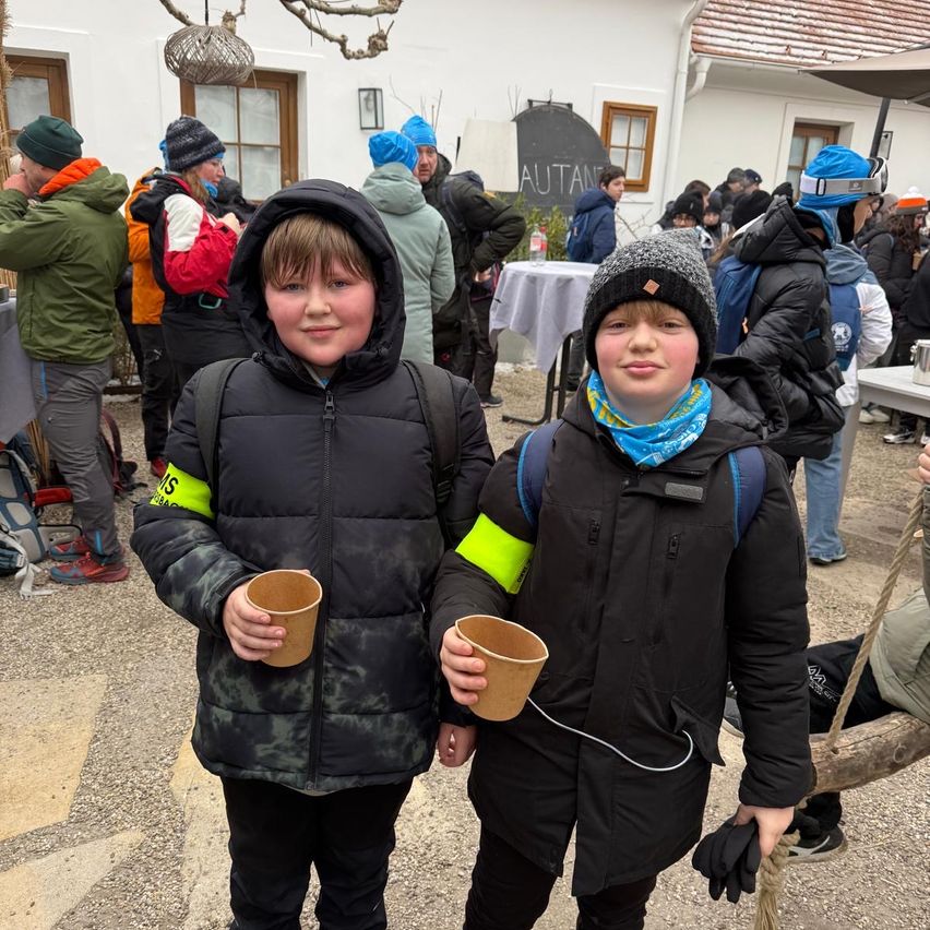 Zwei Jungen in Jacken stehen und halten Tassen in einer verschneiten Außenumgebung mit einer Menge im Hintergrund.