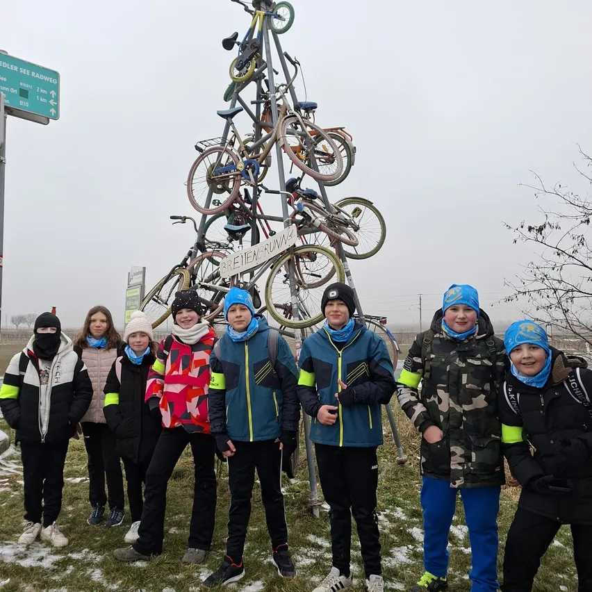Eine Gruppe von Kindern in Winterkleidung posiert vor einem Fahrradbaum. Der Fahrradbaum hat mehrere Fahrräder daran befestigt und steht auf einem Grasfeld.