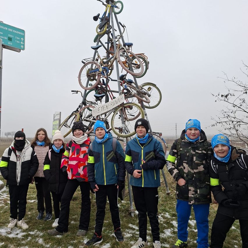 Eine Gruppe von Kindern in Winterkleidung posiert vor einem Fahrradbaum. Der Fahrradbaum hat mehrere Fahrräder daran befestigt und steht auf einem Grasfeld.