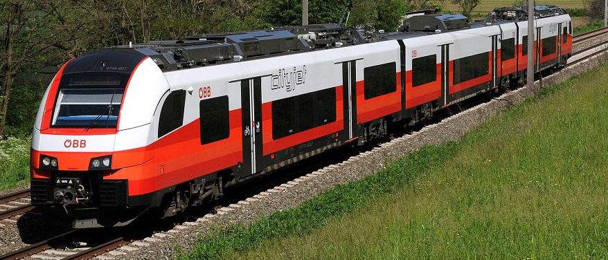 Ein roter und weißer Zug fährt auf einer Strecke in einem ländlichen Gebiet, umgeben von Bäumen und Bergen.