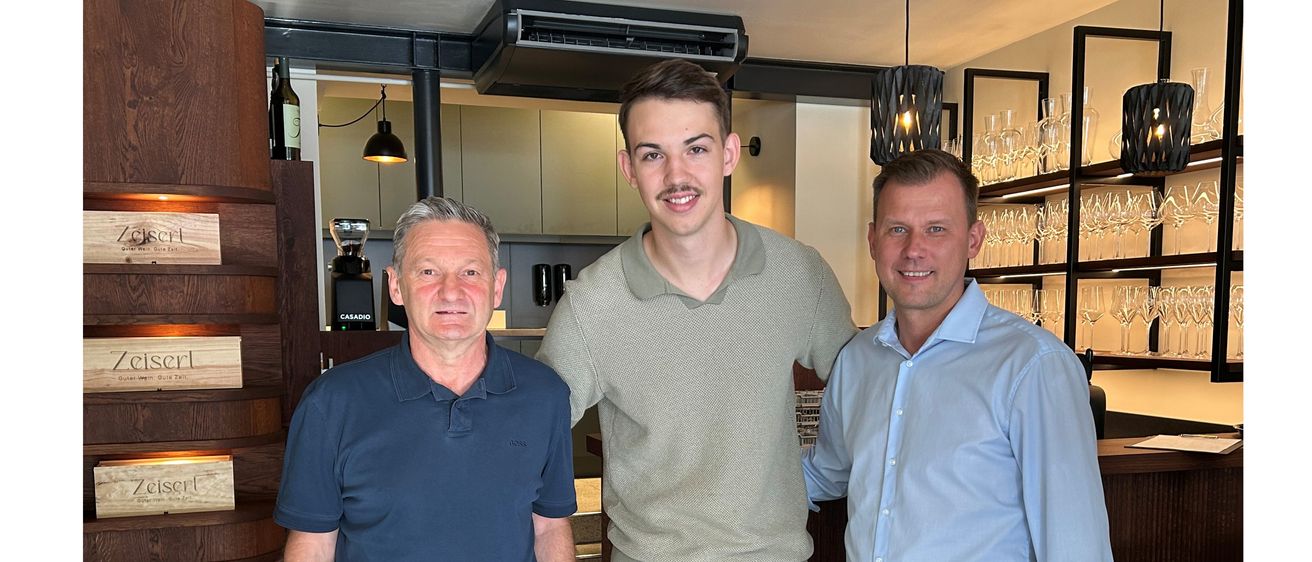 Drei Männer stehen lächelnd in einem Raum zusammen. Der Mann auf der linken Seite trägt ein blaues Poloshirt, der in der Mitte ein graues Hemd und Shorts mit traditionellem Design und der auf der rechten Seite ein hellblaues Langarmhemd. Dahinter befinden sich Regale mit Gläsern.