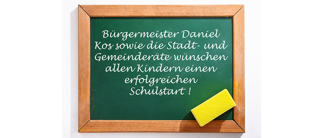 Ein grünes Schultafel mit weißem Text auf Deutsch. Ein gelber Radiergummi befindet sich in der unteren rechten Ecke. Es steht geschrieben: 'Bürgermeister Daniel Kos sowie der Stadt- und Gemeinderat wünschen allen Kindern einen erfolgreichen Schulstart!'