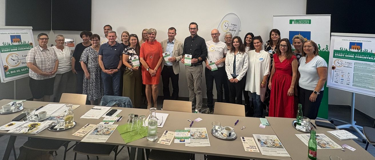 Eine Gruppe von Menschen steht in einem Raum, einige halten Papiere und lächeln. Im Hintergrund befindet sich ein Banner, Monitore und ein Tisch mit Zeitschriften.