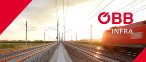 Eine Bahnstrecke mit zwei parallelen Gleisen, umgeben von Strommasten und elektrischen Leitungen. Bäume und Büsche befinden sich an den Seiten der Strecke. Der Himmel ist bewölkt.