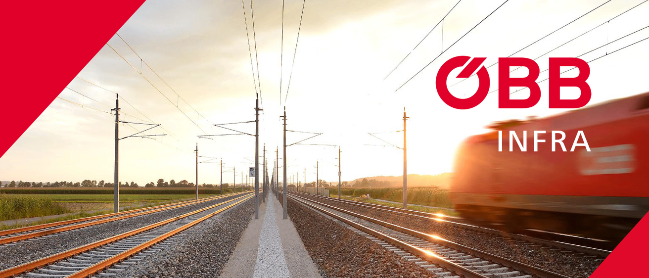 Eine Bahnstrecke mit zwei parallelen Gleisen, umgeben von Strommasten und elektrischen Leitungen. Bäume und Büsche befinden sich an den Seiten der Strecke. Der Himmel ist bewölkt.