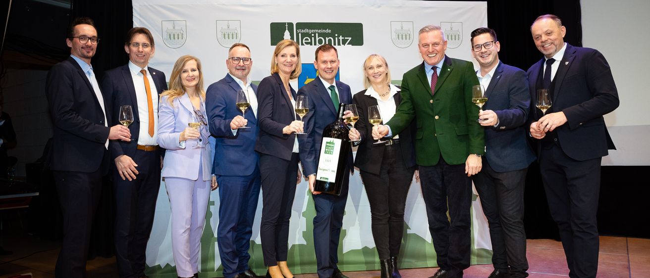 Eine Gruppe formell gekleideter Personen posiert für ein Foto mit Weingläsern und einer Flasche vor einem Banner mit 'Stadtgemeinde Leibnitz' darauf.