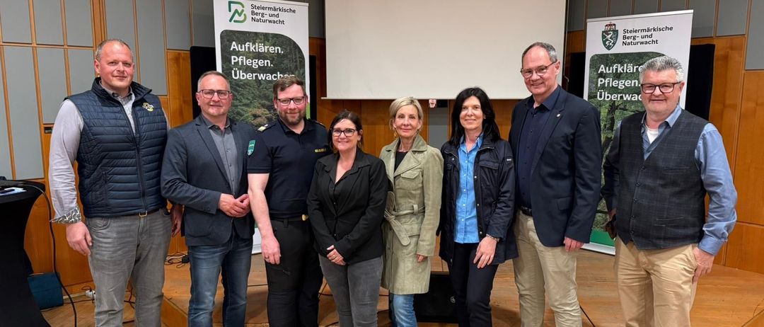 Sieben Personen posieren für ein Foto in einem Konferenzsaal. Sie stehen vor einer Bühne. Drei Banner sind dahinter montiert.