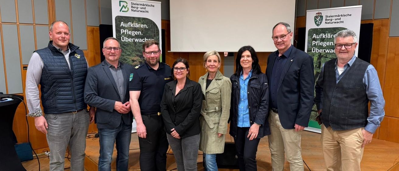 Sieben Personen posieren für ein Foto in einem Konferenzsaal. Sie stehen vor einer Bühne. Drei Banner sind dahinter montiert.