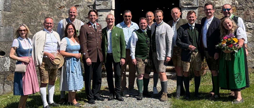 Eine Gruppe von Menschen in traditioneller deutscher Kleidung steht vor einem alten Steingebäude. Der Mann in der Mitte hält einen Hut.