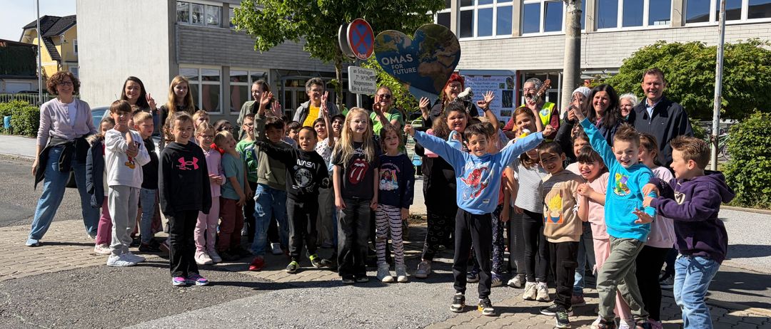 Eine Gruppe von Kindern und Erwachsenen steht auf einem gepflasterten Weg vor einem Gebäude. Die Kinder winken mit den Händen.