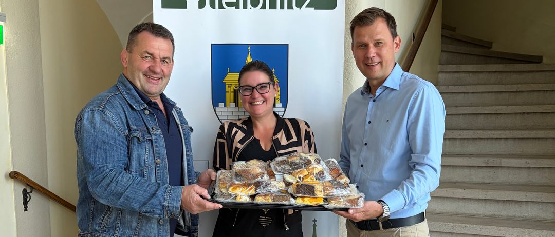 Drei Personen stehen vor einem Banner der Stadt Leibnitz und halten eine Tablett mit Gebäck. Der Mann auf der linken Seite trägt eine Jeansjacke und Jeans, die Frau in der Mitte ein schwarz-weißes Oberteil mit Brille und der Mann auf der rechten Seite ein blaues Hemd und beige Hosen.