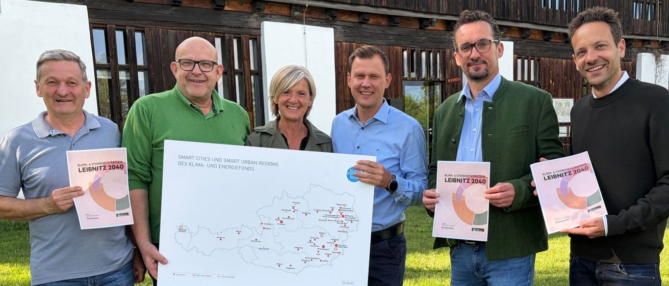 Fünf Personen stehen auf Gras und halten Bücher. Sie lächeln für ein Bild. Die Bücher haben die Worte 'LEIBNITZ 2040'. Sie tragen verschiedene Kleidungsstücke. Dahinter steht ein Gebäude mit Glasfenstern.