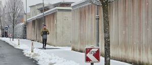 Eine Person joggt auf einem verschneiten Bürgersteig, ein Schild weist auf eine Linkskurve hin. Im Hintergrund befinden sich schneebedeckte Gebäude und ein Baum.