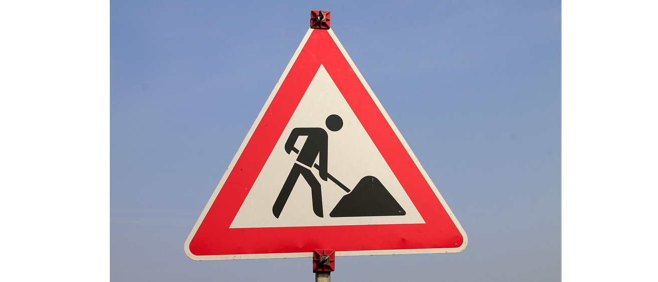 Ein dreieckiges Verkehrsschild mit rotem Rand, weißem Dreieck und schwarzer Silhouette einer Person beim Graben. Das Schild ist an einem Mast gegen einen blauen Himmel montiert.