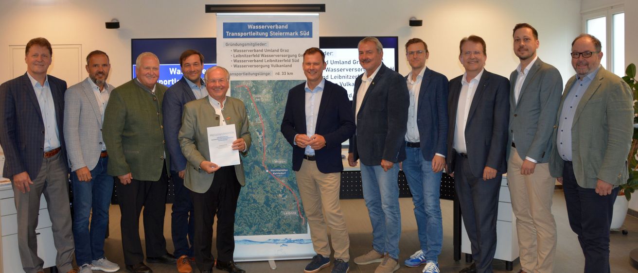 Eine Gruppe von Männern in Geschäftskleidung steht vor einer Karte und hält ein Zertifikat, wahrscheinlich zur Feier eines Projektabschlusses.