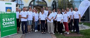 Eine Gruppe von Menschen in weißen T-Shirts steht vor einem Gebäude mit Glasfenstern und einem grünen Banner. Sie lächeln und posieren für ein Foto.