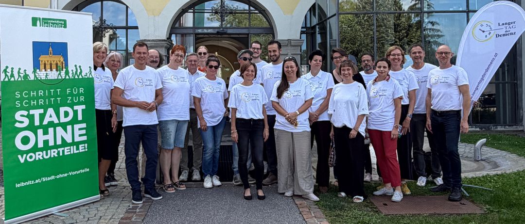 Eine Gruppe von Menschen in weißen T-Shirts steht vor einem Gebäude mit Glasfenstern und einem grünen Banner. Sie lächeln und posieren für ein Foto.
