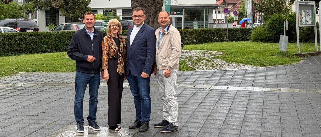 Vier Menschen stehen unter einem großen Baum in einem Park und lächeln. Hinter ihnen befinden sich Gebäude und Grünflächen. Der Boden ist gepflastert und ein Logo ist sichtbar.