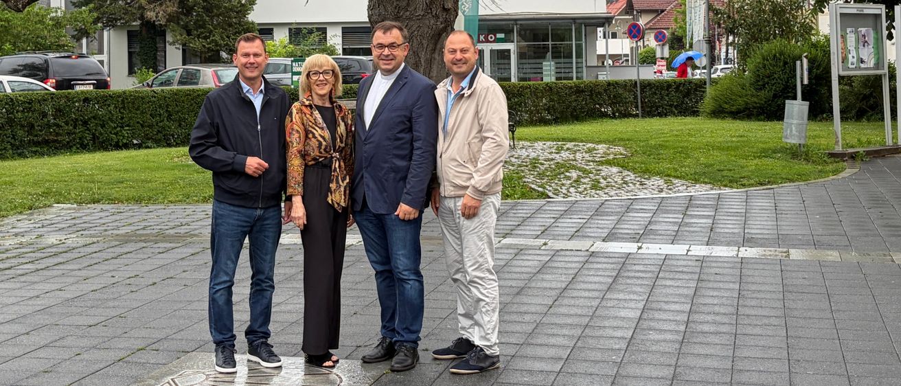 Vier Menschen stehen unter einem großen Baum in einem Park und lächeln. Hinter ihnen befinden sich Gebäude und Grünflächen. Der Boden ist gepflastert und ein Logo ist sichtbar.