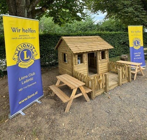 Ein hölzernes Spielhaus mit einem kleinen Tisch und einer Bank steht in einem Hof. Es wird von zwei Bannern mit dem Text 'Lions Club Leibnitz' umrahmt.