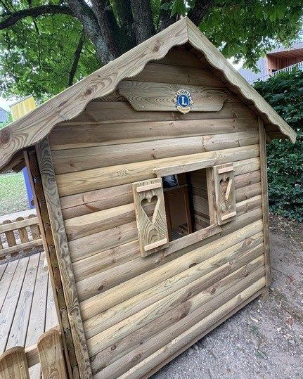 Ein Holzhaus mit einem dreieckigen Dach, einem Fenster und einer Tür. Es befindet sich unter einem Baum in einem Park.