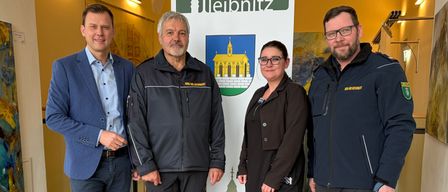 Vier Personen stehen in einem Flur und posieren für ein Foto. Sie alle lächeln. Hinter ihnen befindet sich ein Banner mit den Worten 'stadtgemeinde leipniz' und einem Logo.