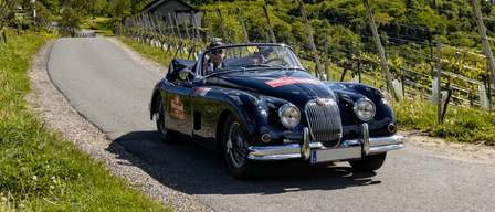 Ein schwarzer Oldtimer fährt auf einer Straße mit einer gelben Kirche und grünen Hügeln im Hintergrund unter einem klaren blauen Himmel.