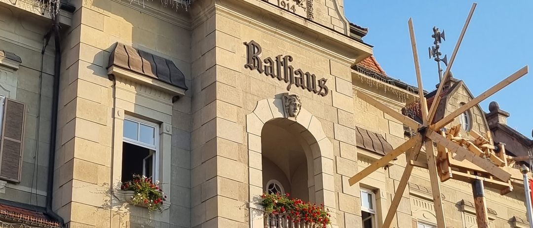 Ein großes Gebäude mit einem Glockenturm, dem Namen Rathaus und mehreren Fahnen davor. Das Gebäude hat viele Fenster und Balkone mit Pflanzen.