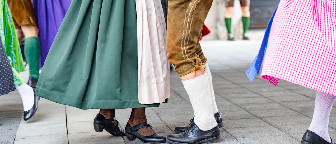 Eine Frau in traditioneller Kleidung tanzt mit einem Mann in Lederhosen auf einem Ziegelboden. Im Hintergrund steht eine weitere Person in traditioneller Kleidung in der Nähe eines Bogens.