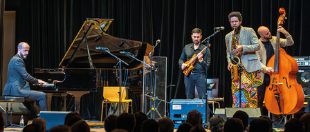 Ein Plakat für ein Jazzfestival mit einer Band, die auf der Bühne auftritt und einem Publikum im Vordergrund. Das Plakat enthält Datum, Uhrzeit und Ort der Veranstaltung sowie eine kurze Beschreibung des Künstlers.