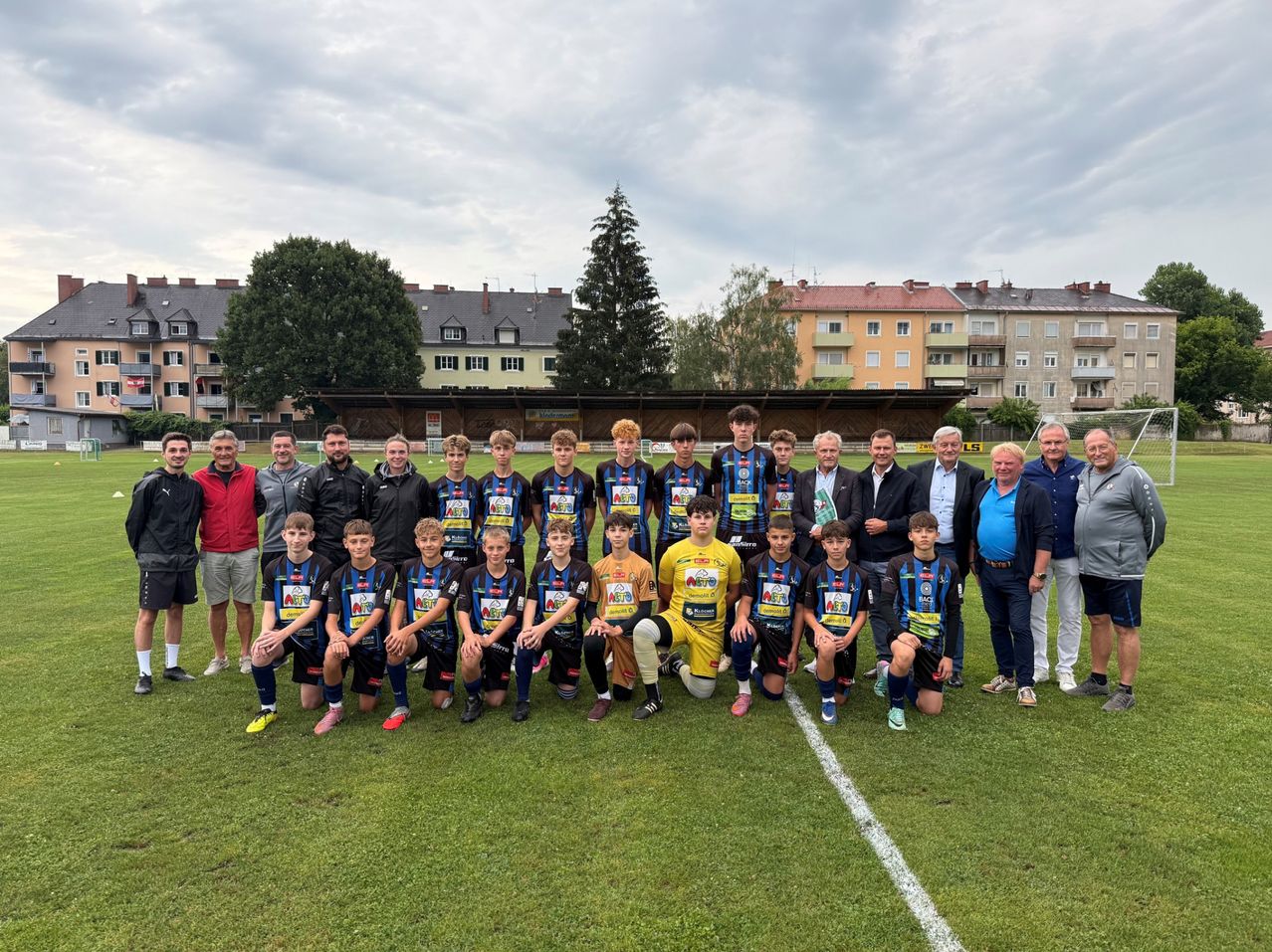 Eine Fußballmannschaft posiert für ein Gruppenfoto auf einem Feld mit Gebäuden und Bäumen im Hintergrund.