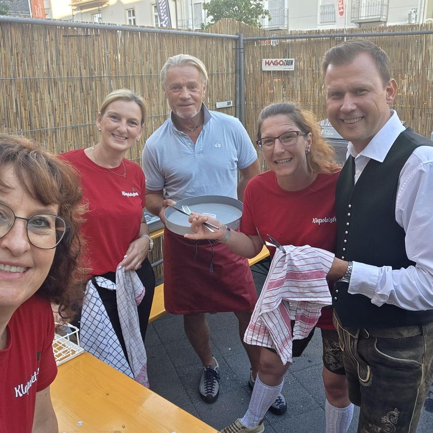 Fünf Menschen lächeln und posieren für ein Foto in einem Außenbereich. Sie tragen rote T-Shirts mit dem Wort 'Klappet' darauf. Der Mann in der Mitte hält einen Teller mit einer Gabel. Hinter ihnen steht ein Bambuszaun mit einem Schild, auf dem 'HAG0ZAUN' steht.