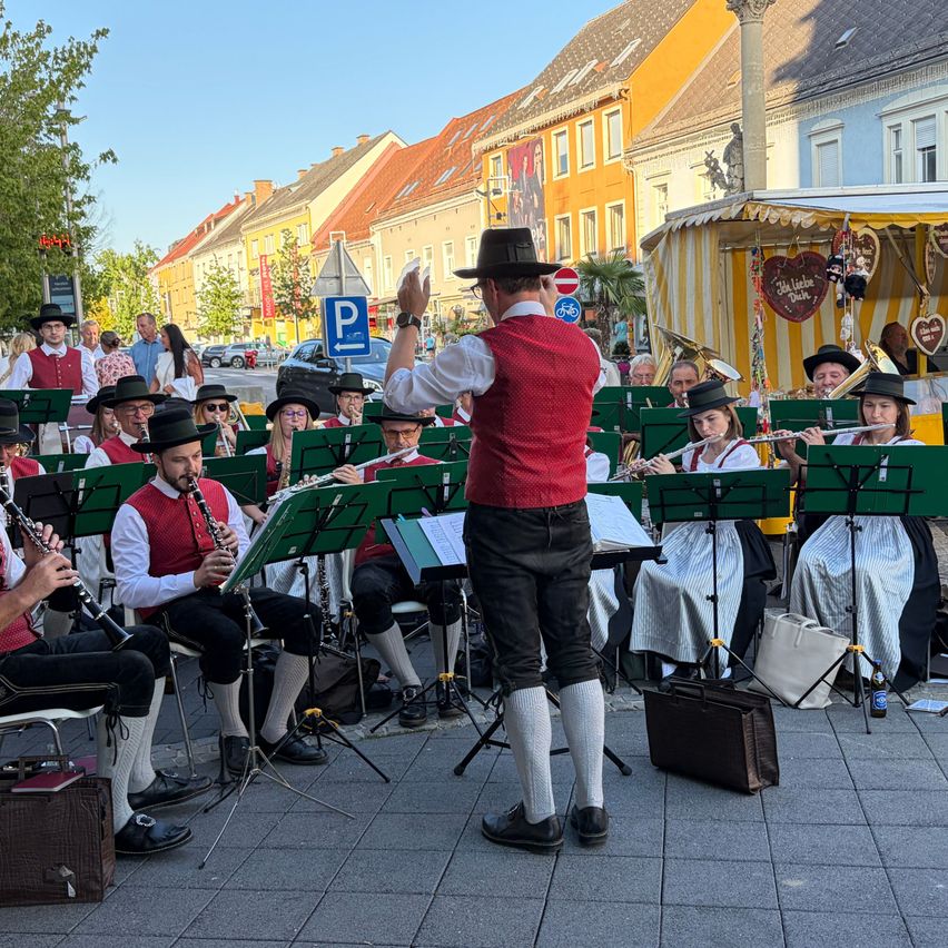 Eine Gruppe von Musikern in traditioneller Kleidung spielt auf einer Stadtstraße, angeführt von einem Dirigenten.