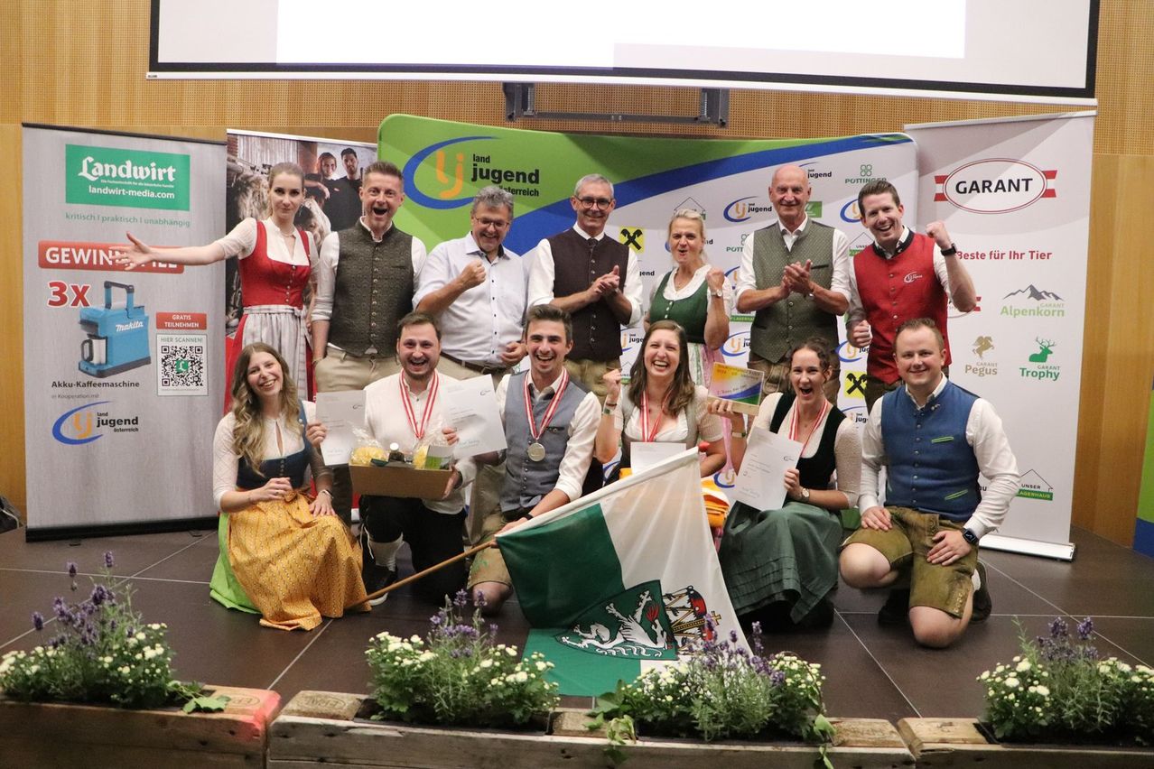 Eine Gruppe von Menschen in traditioneller Kleidung posiert für ein Foto auf der Bühne, hält Urkunden und eine Flagge. Sie lächeln und scheinen eine Leistung zu feiern.