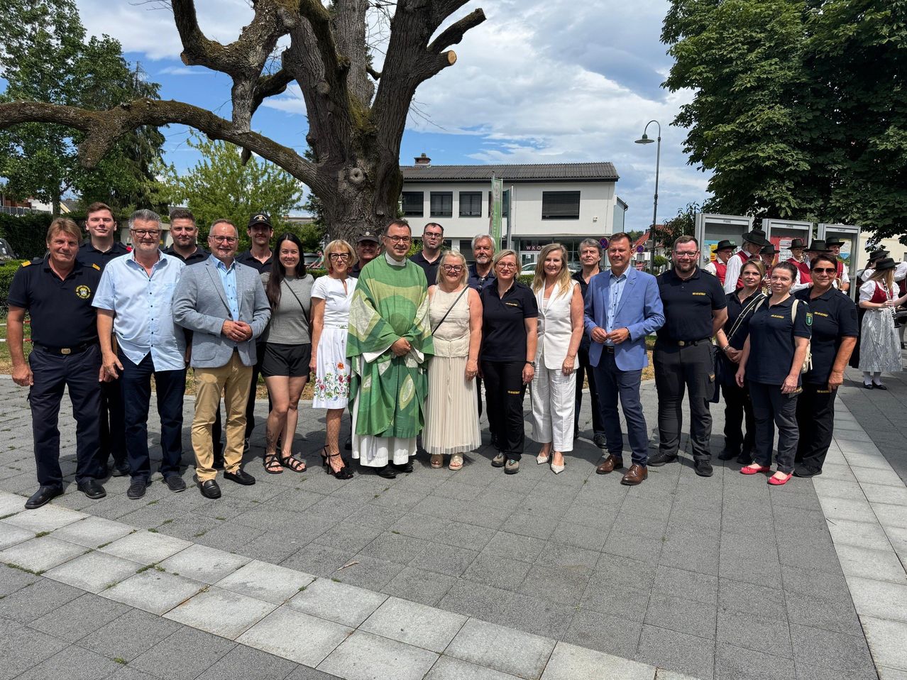 A group of people, including a priest, are standing together in front of a tree and a building on a sunny day.
