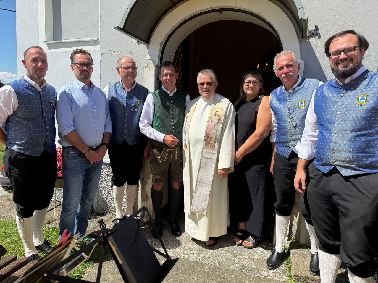 A group of people, including a priest, are standing in front of a church entrance, smiling and posing for a photo.