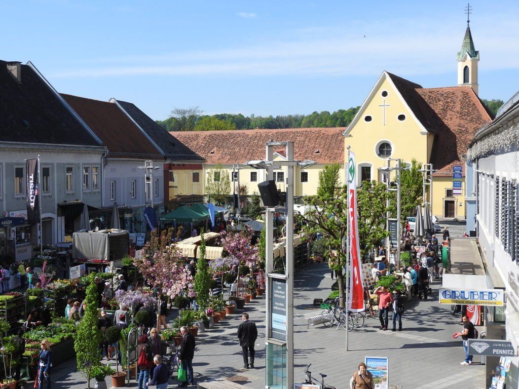 A bustling town square with a church in the background, filled with people shopping and walking around. There are many potted plants and trees, and bicycles are parked on the side.