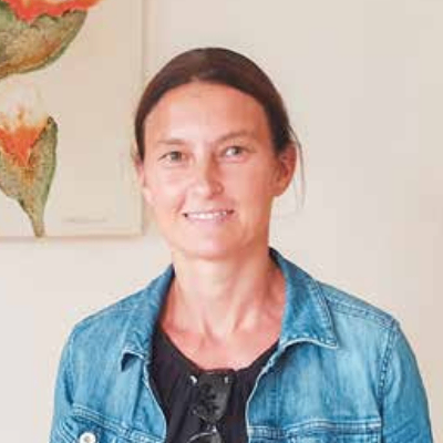 Eine Frau steht vor einer weißen Wand. Sie trägt eine Jeansjacke und ein schwarzes Oberteil. Sie lächelt und posiert für ein Foto.