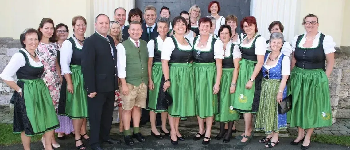 Eine Gruppe von Erwachsenen, darunter Männer und Frauen, die traditionelle deutsche Kleidung tragen, posiert für ein Foto vor einem Gebäude.
