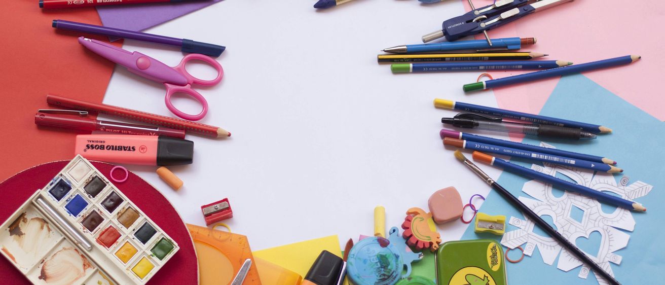 Ein Schreibtisch voller bunter Schulmaterialien. Marker, Scheren, eine Farbpalette und verschiedene Stifte und Bleistifte sind auf einer weißen Oberfläche ausgestellt.